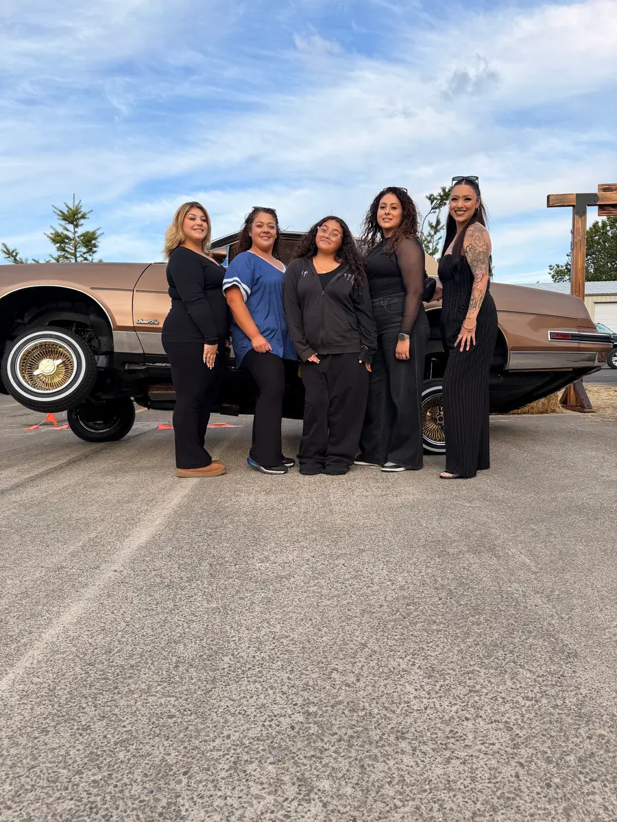 Five women posing in front of a bronze lowrider lifted high off the ground