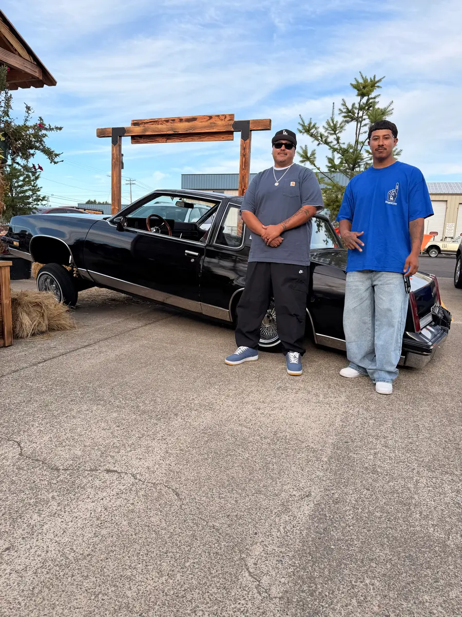 Two men posing beside a black lowrider under a wooden arch